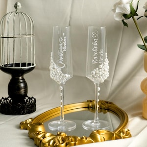 May include: Two clear glass champagne flutes decorated with white faux pearls. The left glass features the name Christian and Nicoleta with a heart icon. The right glass features the name Karolinka and the word Bride with a heart icon.
