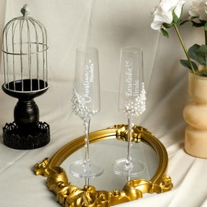 May include: Two clear glass champagne flutes decorated with white pearls. One glass features the white text Christian and Nicoleta with a heart icon, while the other displays Karolinka and Bride with a heart icon. The flutes stand on a gold mirror.