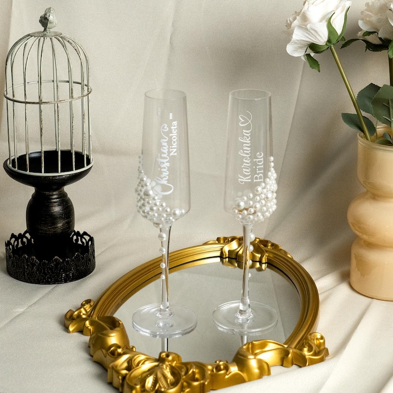 May include: Two clear glass champagne flutes decorated with white pearls. One glass features the white text Christian and Nicoleta with a heart icon, while the other displays Karolinka and Bride with a heart icon. The flutes stand on a gold mirror.