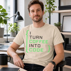 May include: A beige short-sleeved t-shirt featuring the text "I TURN COFFEE INTO CODE" in a mix of black and bright green lettering, framed by bright green curly brackets on the left and right sides of the printed graphic design.