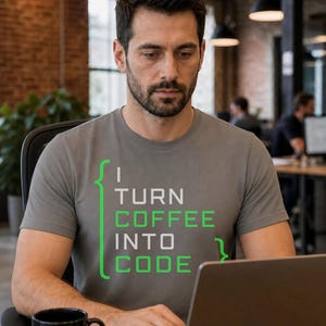 May include: A grey short-sleeved t-shirt featuring the text "I TURN COFFEE INTO CODE" printed in white and green, with green curly brackets framing the text. The shirt has a crew neckline and a standard fit, displayed on a person using a laptop.