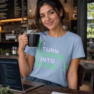 May include: A light blue short-sleeve t-shirt featuring the text "I TURN COFFEE INTO CODE" in white and green lettering, framed by green curly brackets. The shirt is made of a soft fabric and has a crew neckline, providing a casual look.