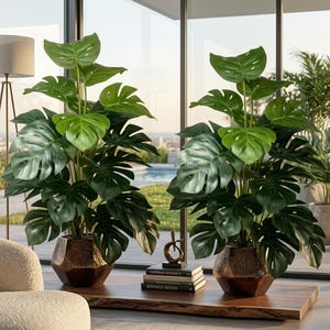 May include: Two artificial monstera plants with large, split green leaves, each potted in a faceted, metallic bronze-coloured planter. The plants are displayed on a wooden surface alongside a stack of three books and a small, abstract gold sculpture.