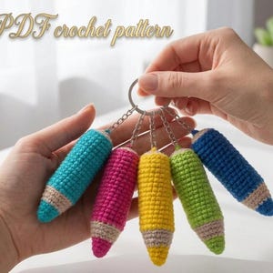May include: A collection of colorful, crocheted pencil keychains in shades of blue, pink, yellow, green, and blue. Each pencil has a beige tip and is attached to a silver keyring. The text "PDF crochet pattern" is visible at the top.