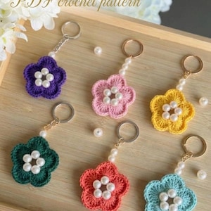 May include: A collection of handmade crochet flower keychains in various colors: purple, pink, yellow, green, red, and blue. Each flower has a pearl center and is attached to a keychain with a pearl bead chain. The text "PDF crochet pattern" is at the top.