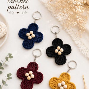 May include: Four crochet flower keychains in navy blue, black, burgundy, and yellow. Each flower has a silver keyring and small wooden beads in the center. The image includes the text "PDF crochet pattern".