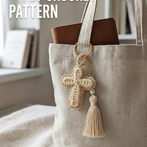 Può includere: Una borsa tote beige con un anello dorato e un portachiavi a forma di croce all'uncinetto. La croce è beige con finiture dorate, una perla e un nappa beige. Il testo "PDF CROCHET PATTERN" è in alto a sinistra. Un portafoglio marrone è dentro la borsa.