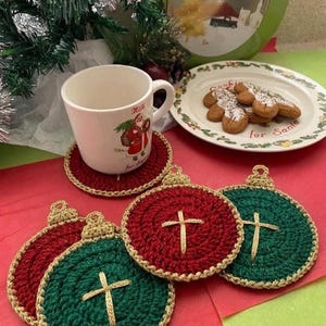 Può includere: Una natura morta a tema natalizio con una tazza bianca con un'illustrazione di Babbo Natale, biscotti di pan di zenzero e sottobicchieri all'uncinetto a forma di ornamento in rosso e verde. Una scatola di Natale e del verde completano la scena.
