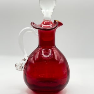 May include: A ruby red glass cruet with a clear glass stopper and handle. The cruet has a bulbous body and a flared rim. The stopper is elongated and rounded. The handle is curved and attached to the side of the cruet. Decorative glassware.
