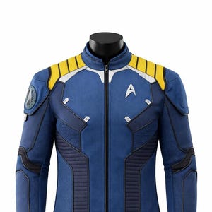 Può includere: Giacca blu con dettagli gialli sulle spalle e cerniera argentata. La giacca presenta un'insegna di Starfleet e altri dettagli. La giacca è esposta su un manichino nero.