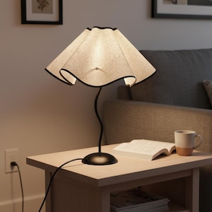 May include: A table lamp featuring a beige, textured, wavy fabric shade with a thin black trim. The lamp has a slender, curved black metal stem and a circular black base. The light emits a warm glow, illuminating the shade's fabric texture.