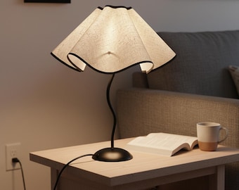 May include: A table lamp featuring a beige, textured, wavy fabric shade with a thin black trim. The lamp has a slender, curved black metal stem and a circular black base. The light emits a warm glow, illuminating the shade's fabric texture.
