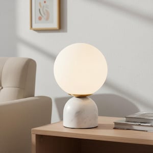 May include: A modern table lamp with a white globe shade and a marble base. The lamp has a gold-colored accent and sits on a wooden surface. The lamp is turned on, emitting a soft glow.