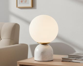 May include: A modern table lamp with a white globe shade and a marble base. The lamp has a gold-colored accent and sits on a wooden surface. The lamp is turned on, emitting a soft glow.