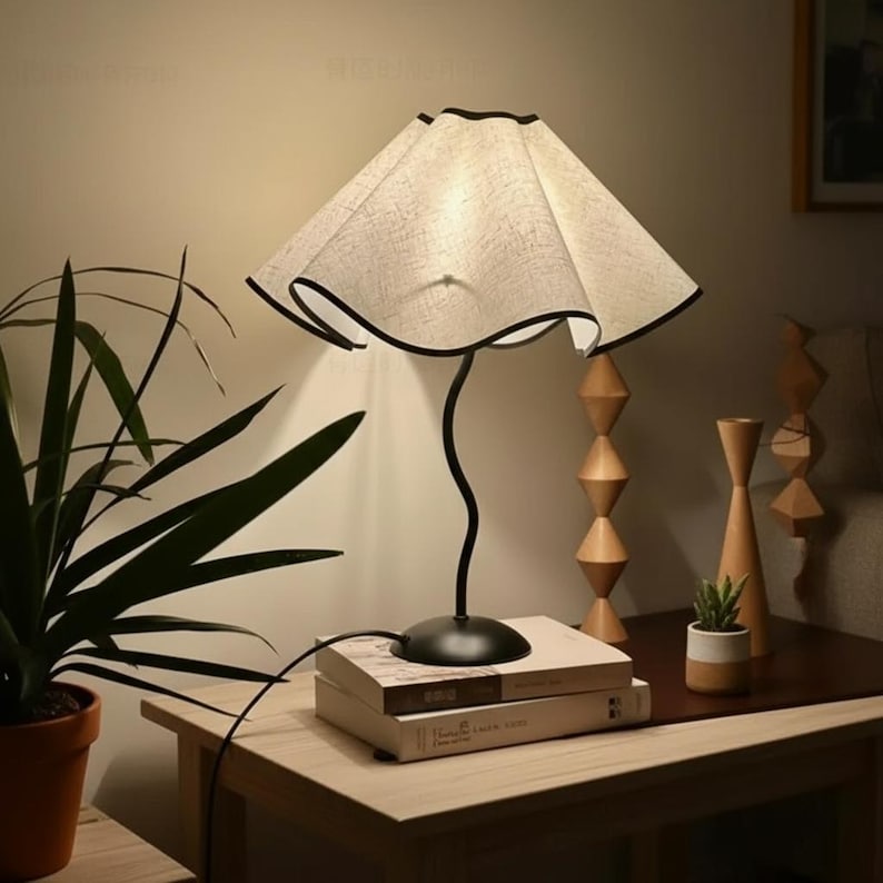 May include: A table lamp with a black, wavy metal stem and a wide, beige, fabric-covered shade featuring a scalloped edge with a thin black trim. The lamp base is circular and black, resting on a stack of two books on a wooden table surface.