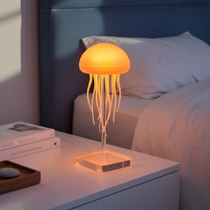 Peut inclure: Lampe en forme de méduse avec une lueur orange chaude. La lampe a une base carrée transparente et une tige transparente soutenant le design de la méduse. Le corps de la méduse est un dôme arrondi, avec des tentacules suspendues. Elle est posée sur une table de chevet blanche.