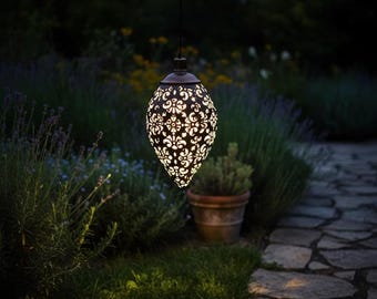 May include: A teardrop-shaped hanging lantern with a dark bronze finish and intricate laser-cut floral patterns. The metal structure emits a warm, glowing light from within, casting detailed shadows through the cutouts in the dark outdoor setting.