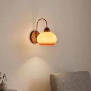 Peut inclure: Une applique murale composée d'une base circulaire en bois, d'un bras courbé en laiton et d'un abat-jour arrondi en verre dépoli diffusant une lueur douce et chaude. Le luminaire est fixé au mur et projette une lumière vers le bas.
