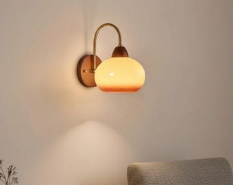 May include: A wall-mounted lamp featuring a circular wooden base, a curved brass arm, and a rounded, frosted glass shade that emits a warm, soft glow. The fixture is attached to a plain wall, providing downward illumination in a room.