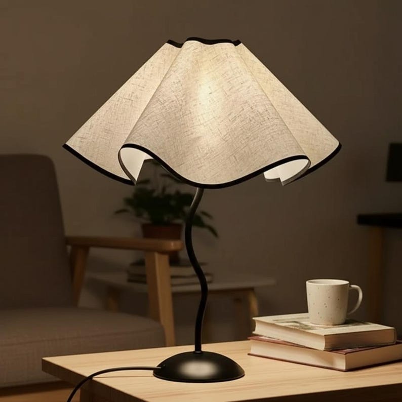 May include: A table lamp featuring a beige, textured, wavy fabric lampshade with a thin black trim. The lamp has a slender, curved black metal stem and a circular black base, providing a soft, warm glow from the illuminated shade.