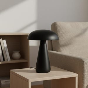 Puede incluir: Una lámpara de mesa negra con forma de hongo, con una parte superior redondeada y una base cónica. La lámpara está hecha de un material negro mate y se encuentra sobre una mesita auxiliar de madera clara. La lámpara está enchufada.