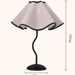 May include: A table lamp with a wavy, beige fabric shade featuring a black trim. The lamp has a slender, curved black metal stem and a round black base. The total height is 390mm and the base diameter is 120mm as indicated by the measurement lines.