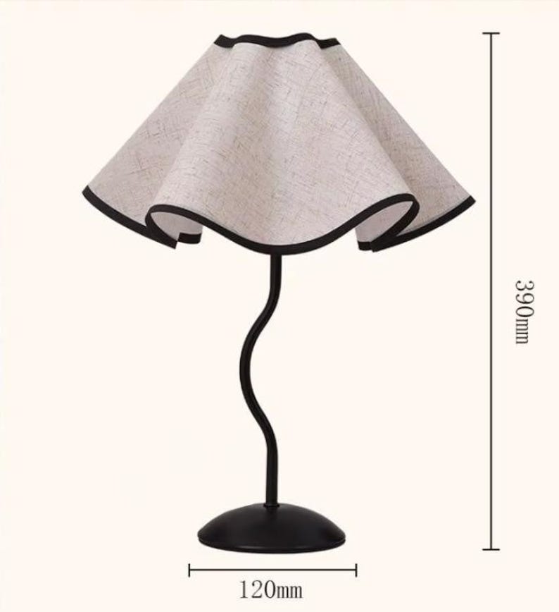 May include: A table lamp with a wavy, beige fabric shade featuring a black trim. The lamp has a slender, curved black metal stem and a round black base. The total height is 390mm and the base diameter is 120mm as indicated by the measurement lines.