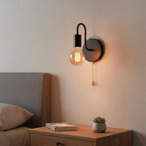 May include: Black wall-mounted light fixture with a round backplate and a curved arm holding a round, exposed light bulb. A pull chain with a wooden toggle hangs down. The fixture is mounted on a light-colored wall.