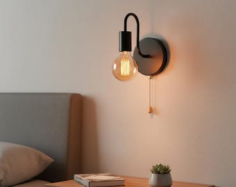 May include: Black wall-mounted light fixture with a round backplate and a curved arm holding a round, exposed light bulb. A pull chain with a wooden toggle hangs down. The fixture is mounted on a light-colored wall.