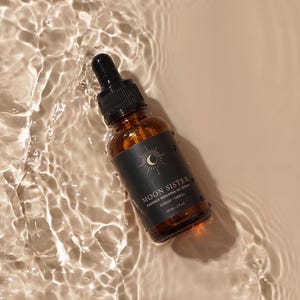 May include: A brown glass bottle with a black dropper, labeled "Moon Sister Radiance Renewing Oil Serum." The bottle is partially submerged in water, creating ripples. The label features a sun and moon design and the text "Rosehip + Neroli."