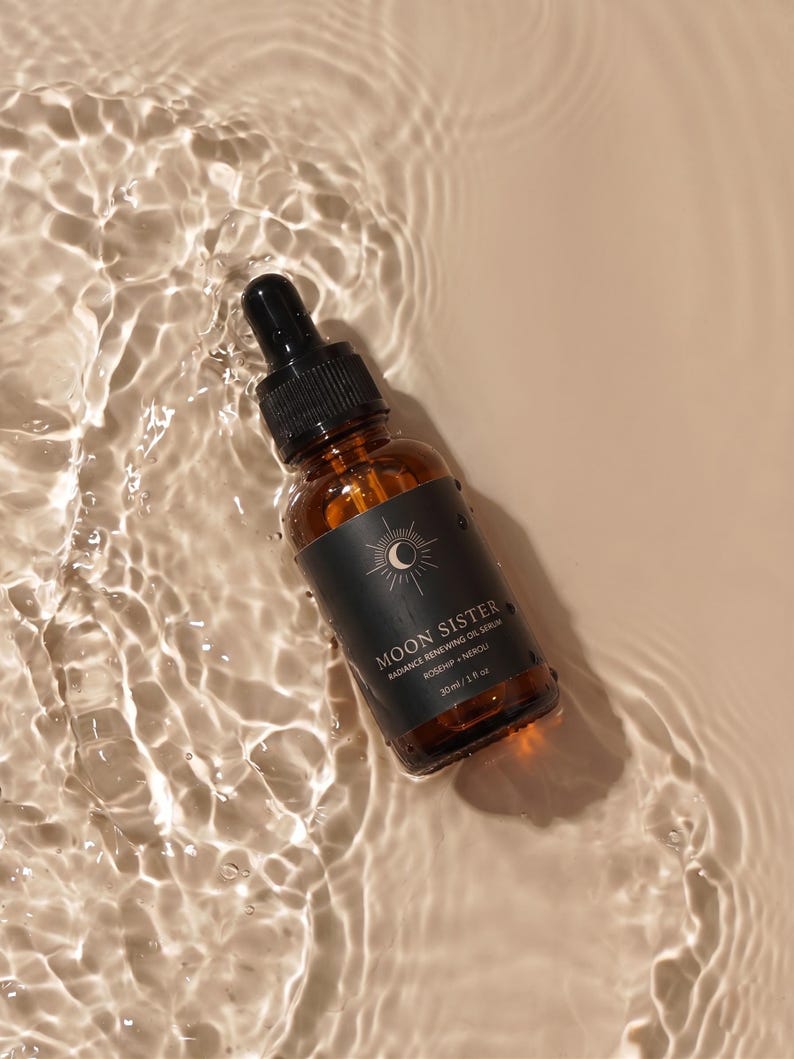 May include: A brown glass bottle with a black dropper, labeled "Moon Sister Radiance Renewing Oil Serum." The bottle is partially submerged in water, creating ripples. The label features a sun and moon design and the text "Rosehip + Neroli."