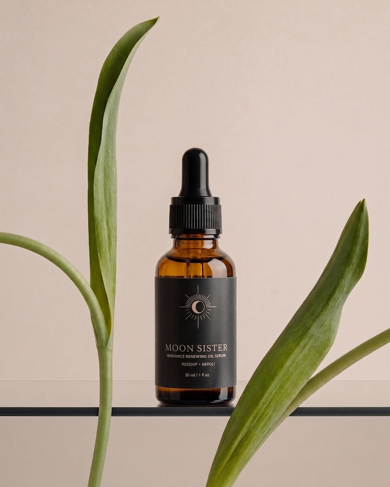 May include: A brown glass bottle with a black dropper and a black label. The label reads "MOON SISTER" and "RADIANCE RENEWING OIL SERUM". The bottle is 1 fl oz (30 ml). Green leaves frame the bottle against a neutral background.