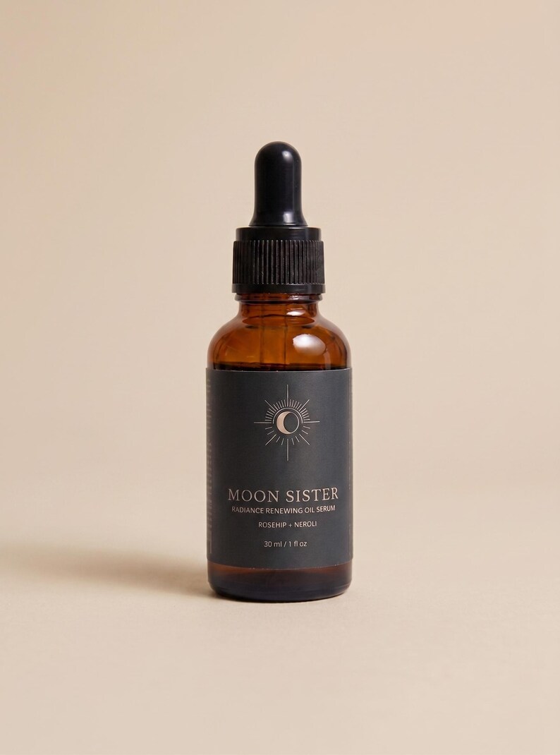 May include: A brown glass bottle of Moon Sister Radiance Renewing Oil Serum with a black label. The label features a sun and moon design and the text "Rosehip + Neroli". The bottle contains 1 fl oz of serum.