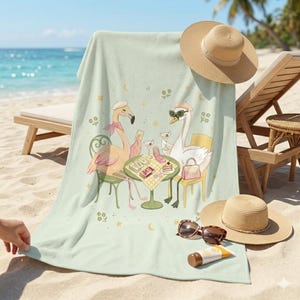 May include: A light green beach towel featuring a whimsical illustration of a flamingo and a swan enjoying cocktails at a table. The towel is decorated with stars, moons, and floral accents. A straw hat and sunglasses are also visible.