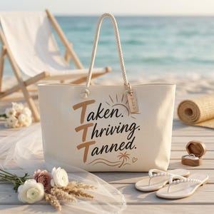 May include: A cream-colored canvas tote bag with rope handles and a wooden tag labeled Bride to Be. The bag features the brown and black text Taken. Thriving. Tanned. along with a sun, palm tree, and heart graphic printed on the front surface.