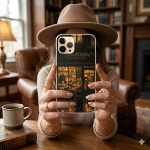 May include: A phone case featuring a detailed illustration of a bakery storefront with the text "Moonlight Bakery". The case has a dark color scheme with warm, inviting interior lighting. The phone is held in front of a person wearing a hat.