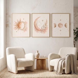Puede incluir: Tres impresiones artísticas enmarcadas con diseños celestiales. Las impresiones presentan un sol, una luna creciente con el perfil de una mujer y fases lunares, todo en tonos melocotón y dorado. La obra de arte cuelga sobre dos sillones color crema.