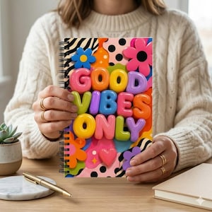 May include: A spiral-bound notebook with the words "GOOD VIBES ONLY" in colourful, 3D bubble letters. The cover features a vibrant design with flowers, hearts, and abstract shapes in pink, orange, blue, and purple. The notebook is held by a person.