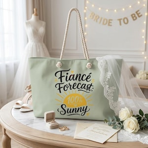 May include: A sage green tote bag with rope handles featuring the black text Fiance Forecast: 100% Sunny and a yellow sun graphic. The bag is displayed with a white veil, a ring box containing a ring, two gold rings, and a wedding invitation card.