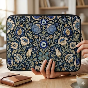 May include: A laptop sleeve with a dark blue floral pattern featuring blue and cream-colored flowers, pomegranates, and evil eye designs. The sleeve has a black zipper and is held on a wooden table.