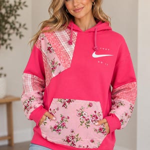 May include: A bright pink hooded sweatshirt featuring a white Nike swoosh logo and the text JUST DO IT. The garment includes a front kangaroo pocket and sleeves accented with light pink floral and plaid patchwork fabric panels.