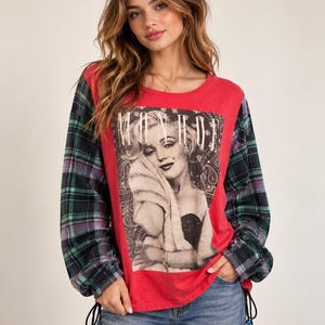 Puede incluir: Una camisa de manga larga con un panel frontal rojo que presenta un gráfico en blanco y negro de Marilyn Monroe y el texto MONROE. Las mangas son de tela a cuadros verde, negra y morada. El dobladillo tiene cordones negros ajustables con cuentas azules.