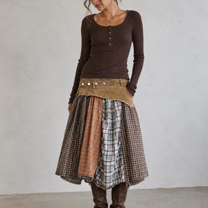 May include: A dark brown long-sleeve ribbed henley shirt with a scoop neckline and buttons, paired with a multi-patterned plaid midi skirt featuring a tan denim-style waistband and brown leather knee-high boots with side buckles.