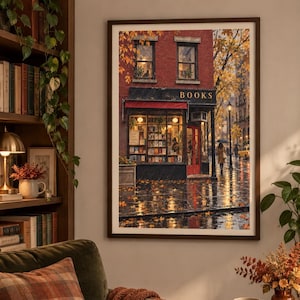 May include: A framed wall art print depicting a rainy city street scene with a brick bookstore building. The storefront features a red awning, a red door, and large windows displaying shelves of books. Golden autumn leaves cover the wet pavement.