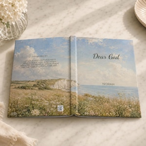 May include: An open book with a landscape painting on the cover and pages. The left side features text from Proverbs 3:3. The right side reads "Dear God" with the name "Norah" below. The book is on a marble surface.