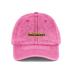May include: A pink, vintage-style baseball cap with a curved brim. The cap features the word "hellraiser" embroidered in yellow script across the front. The hat has a slightly faded appearance.