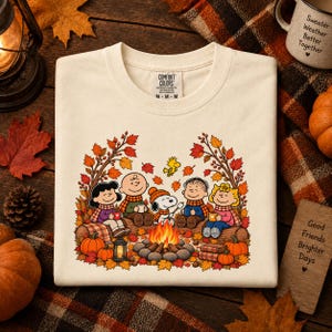 May include: A cream-colored t-shirt featuring a graphic of the Peanuts characters sitting around a campfire. The design includes Snoopy, Charlie Brown, Lucy, Linus, and Sally surrounded by autumn leaves, pumpkins, and a lantern. The shirt has a Comfort Colors brand label at the neck.