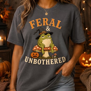 May include: A charcoal gray short-sleeve t-shirt featuring a graphic of a green frog wearing a black witch hat, sitting on a red and white spotted mushroom. The text "FERAL & UNBOTHERED" is printed in orange and cream above and below the frog.