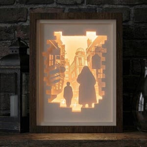 May include: A framed, illuminated paper art piece depicting a detailed street scene with figures and buildings. The artwork is encased in a wooden frame, with the light emanating from within, creating a warm, inviting glow. The scene is a silhouette of a city street.