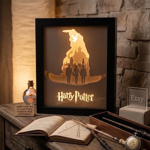Könnte beinhalten: Ein schwarz gerahmter Harry Potter Schattenkasten mit einem geschichteten Design, das den sprechenden Hut, Silhouetten von Charakteren und den Text "Harry Potter" zeigt. Die Box ist beleuchtet und erzeugt ein warmes Leuchten.